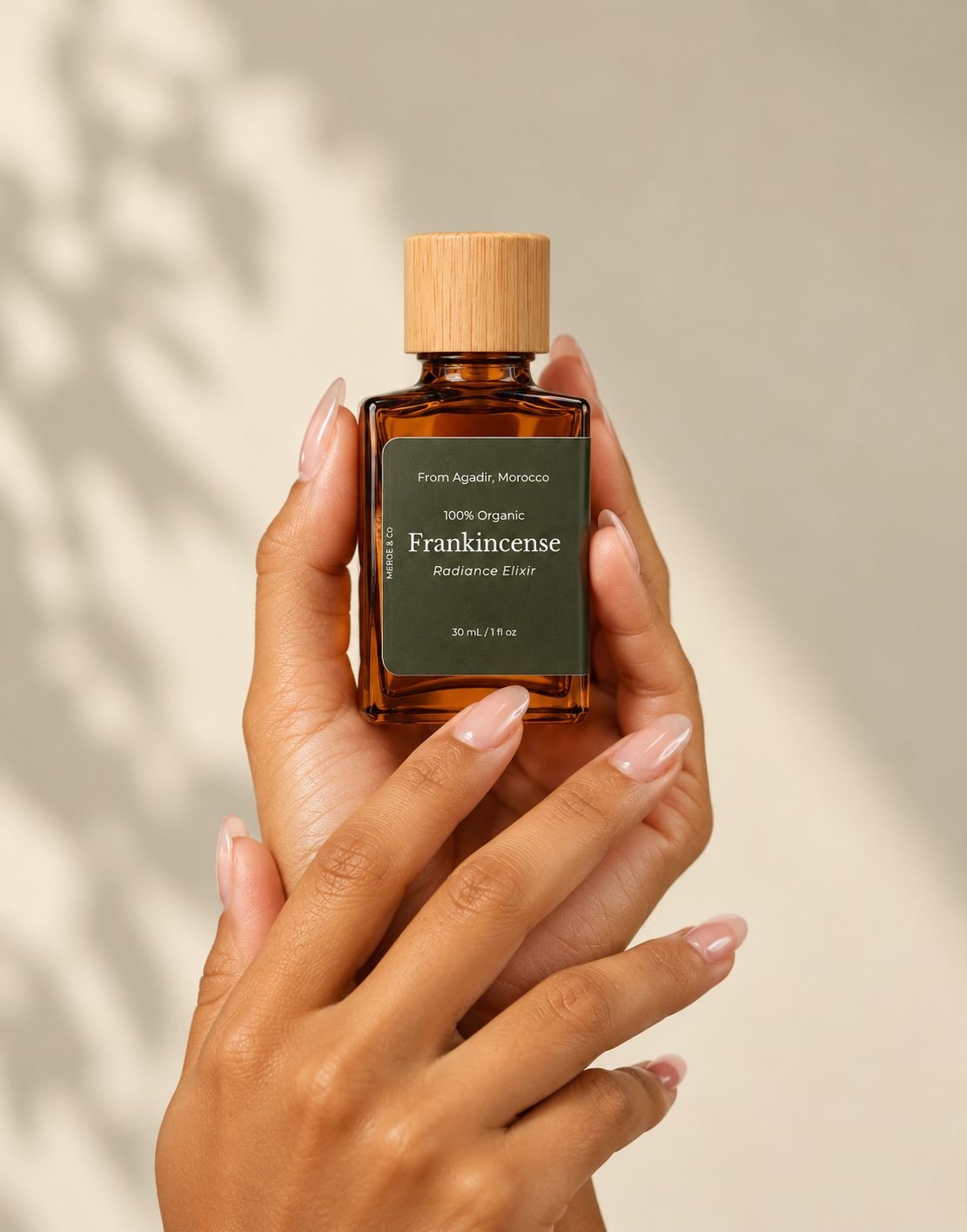 Hands holding a MEROË Frankincense elixir in soft leaf-shadow light